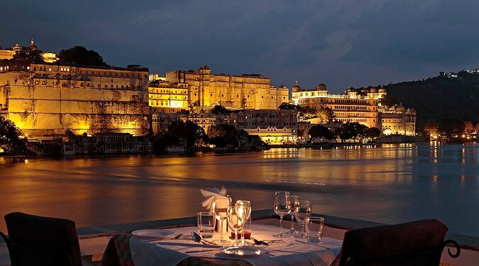 A romantic evening view from a restaurant with a set table overlooking the illuminated palaces of Udaipur across a lake.