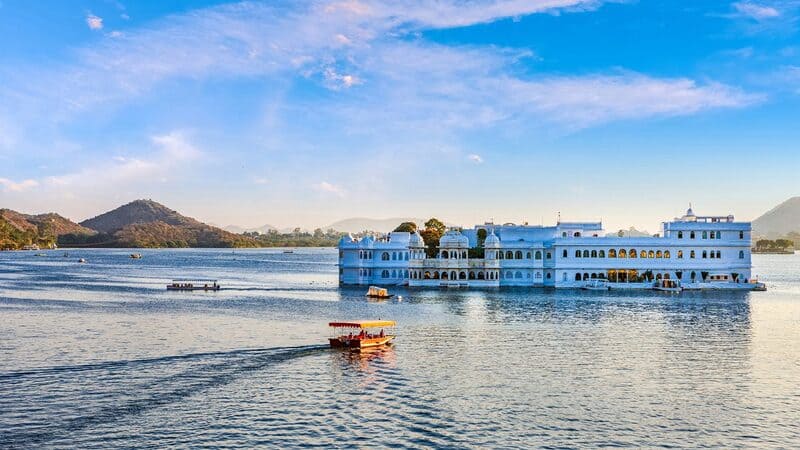 White Taj Lake Palace situated in the middle of Lake Pichola with boats sailing across the sparkling water and Aravalli hills in the background. 