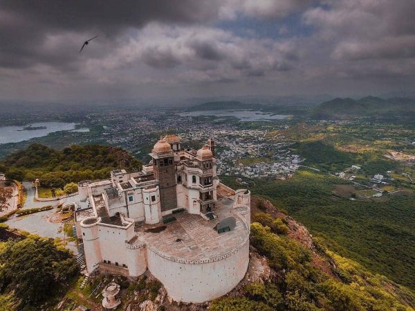 Aerial shot of Monsoon Palace perched atop a hill with a panoramic view of Udaipur city and lakes under a dramatic cloudy sky.
